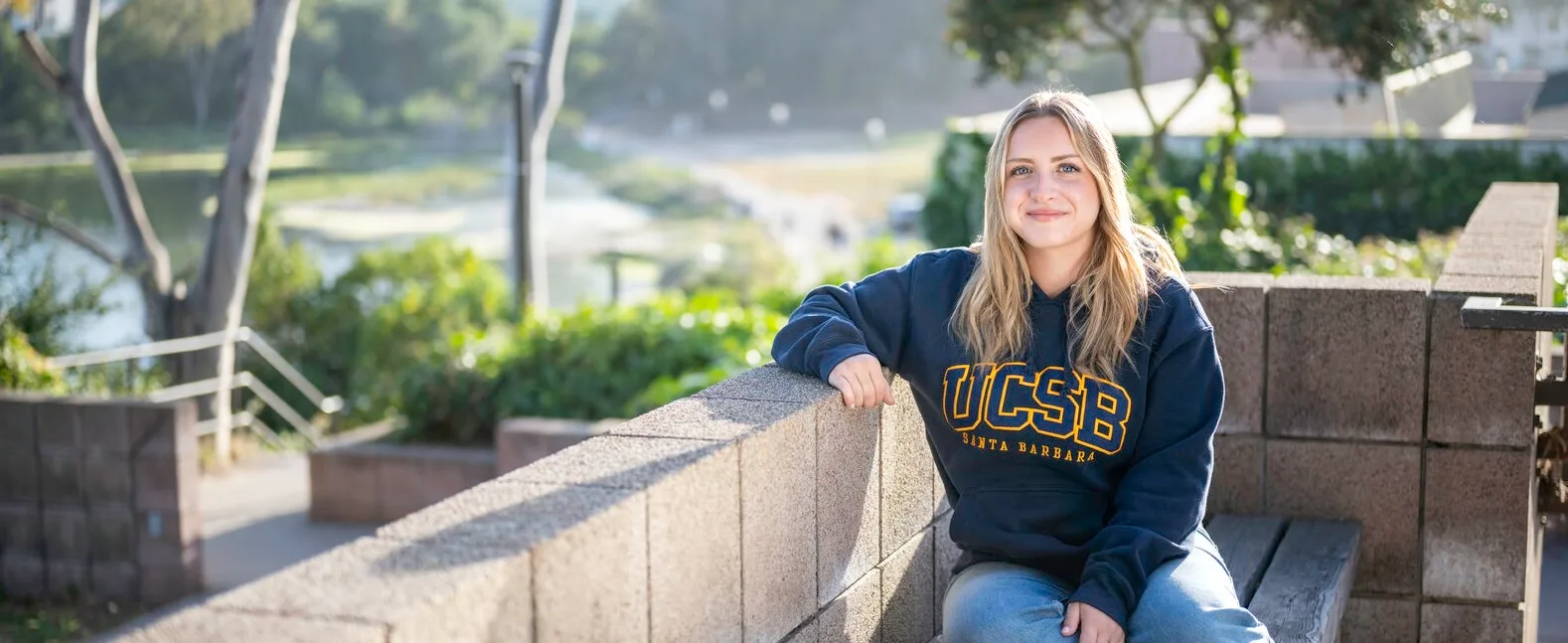 Woman in a UCSB sweatshirt seated on an outdoor bench beside a low stone wall